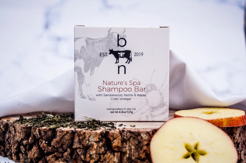 May include: A white box of Nature's Spa Shampoo Bar with a cow and bee logo, and the text "EST. 2019". The box is on a wooden surface with tea leaves and apple slices. The net weight is 4.4oz/125g.