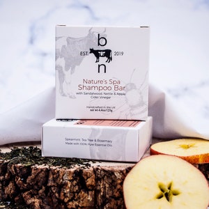May include: Two white boxes of shampoo bars with text, one labeled "Nature's Spa Shampoo Bar" with sandalwood, nettle, and apple cider vinegar. The other box is labeled "Spearmint, Tea Tree & Rosemary". The boxes are on a wooden surface with apple slices and tea leaves.