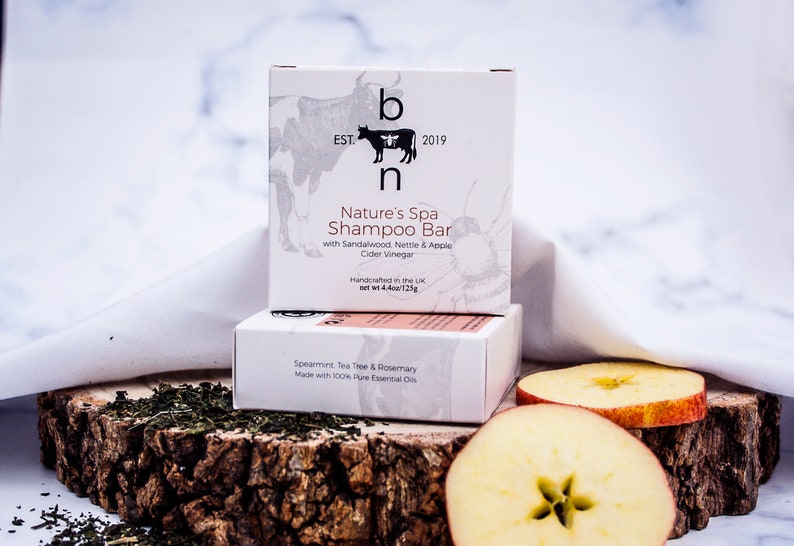 May include: Two white boxes of shampoo bars with text, one labeled "Nature's Spa Shampoo Bar" with sandalwood, nettle, and apple cider vinegar. The other box is labeled "Spearmint, Tea Tree & Rosemary". The boxes are on a wooden surface with apple slices and tea leaves.
