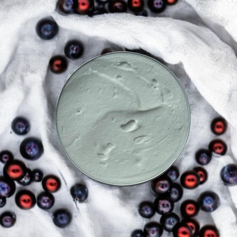May include: A close-up of a light blue, whipped, creamy substance in a white container. The container is surrounded by blueberries and red berries on a white fabric background.