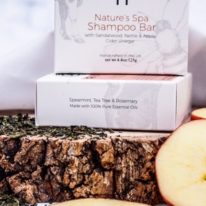 May include: Two white boxes of shampoo bars are stacked on a wooden surface, with text that reads "Nature's Spa Shampoo Bar" and "Spearmint, Tea Tree & Rosemary." The image includes dried herbs and apple slices. The top box is labeled "Handcrafted in the UK net wt 4.4oz/125g."