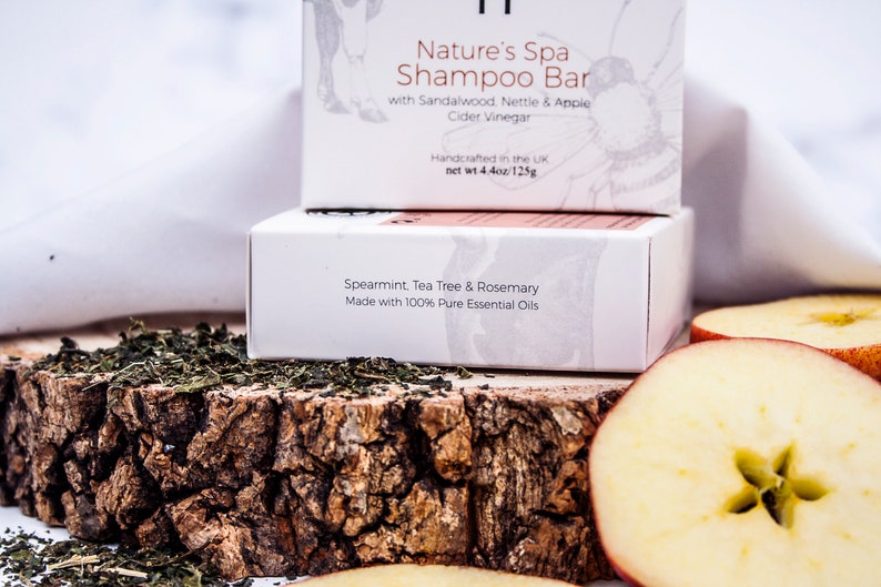 May include: Two white boxes of shampoo bars are stacked on a wooden surface, with text that reads "Nature's Spa Shampoo Bar" and "Spearmint, Tea Tree & Rosemary." The image includes dried herbs and apple slices. The top box is labeled "Handcrafted in the UK net wt 4.4oz/125g."
