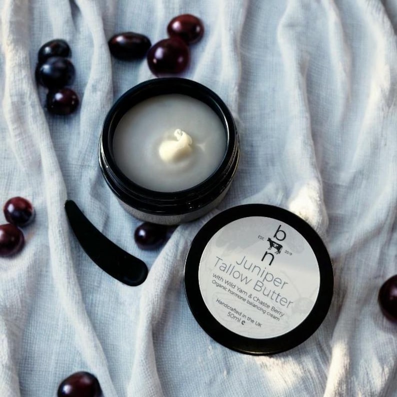 May include: A black jar with a white label that reads "Juniper Tallow Butter with Wild Yam & Chaste Berry Organic hormone balancing cream Handcrafted in the UK 50ml e". The jar is open and contains a white cream.