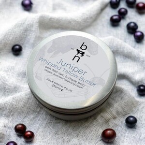 May include: A silver tin of Juniper Whipped Tallow Butter with Wild Yam & Chaste Berry. The tin is labeled with the brand name "bh" and the text "EST. 2019". The tin is sitting on a white fabric background with dark blue berries scattered around it.