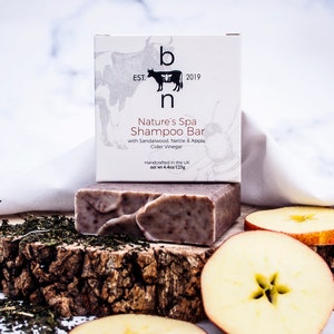 May include: A shampoo bar and its packaging are displayed on a wooden surface with apple slices and tea leaves. The packaging reads "Nature's Spa Shampoo Bar" with sandalwood, nettle, and apple cider vinegar. The bar weighs 4.4 oz / 125g.