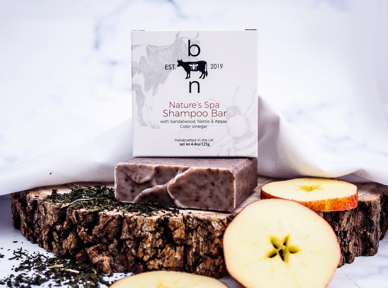 May include: A shampoo bar and its packaging are displayed on a wooden surface with apple slices and tea leaves. The packaging reads "Nature's Spa Shampoo Bar" with sandalwood, nettle, and apple cider vinegar. The bar weighs 4.4 oz / 125g.