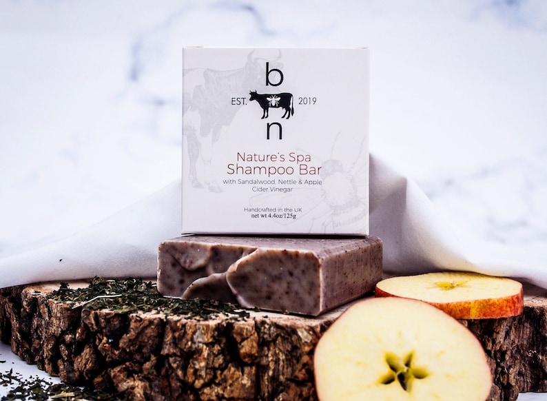 May include: A shampoo bar and its box are displayed on a wooden surface with apple slices. The box reads "Nature's Spa Shampoo Bar" with sandalwood, nettle, and apple cider vinegar. The bar is 4.4 oz.