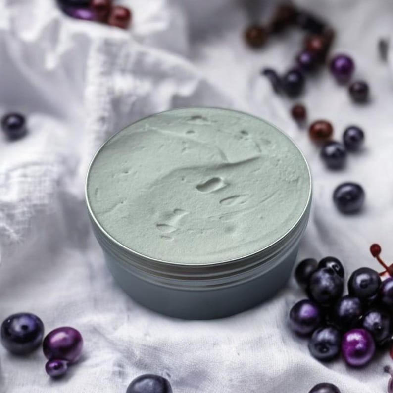 May include: A close-up of a light blue, round container filled with a light green, whipped cream-like substance. The container is sitting on a white fabric surface with dark purple berries scattered around it.