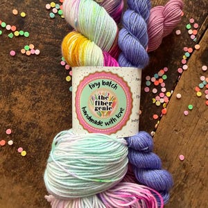 May include: A collection of colorful yarn skeins in shades of pink, yellow, blue, and purple, with a decorative pin. The yarn is arranged on a wooden surface with confetti. A label reads "tiny batch the fiber genie handmade with love."