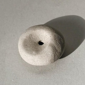 Puede incluir: Un peque&ntilde;o objeto de piedra gris claro en forma de rosquilla con un agujero central. La piedra tiene una superficie texturizada y proyecta una sombra sobre un fondo gris. El objeto parece hecho a mano.
