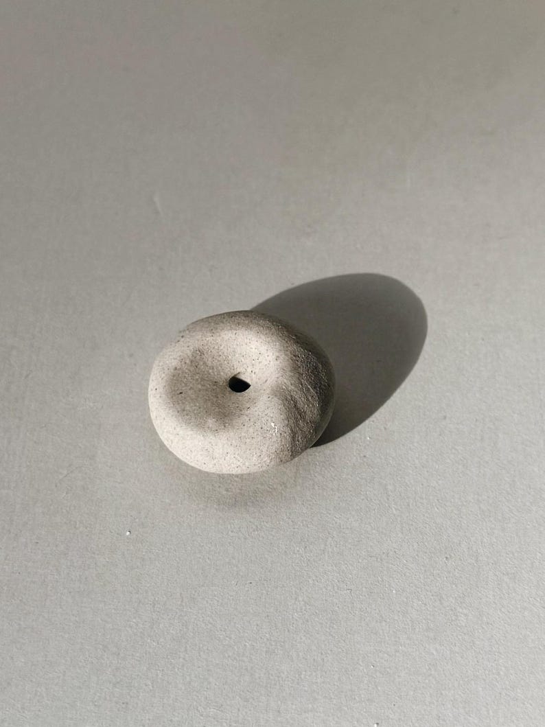 Puede incluir: Un peque&ntilde;o objeto de piedra gris claro en forma de rosquilla con un agujero central. La piedra tiene una superficie texturizada y proyecta una sombra sobre un fondo gris. El objeto parece hecho a mano.