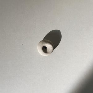Puede incluir: Una peque&ntilde;a cuenta de cer&aacute;mica blanquecina con forma circular y un agujero central. La cuenta es lisa y tiene un acabado mate. La imagen est&aacute; tomada desde arriba, con una sombra proyectada hacia un lado.