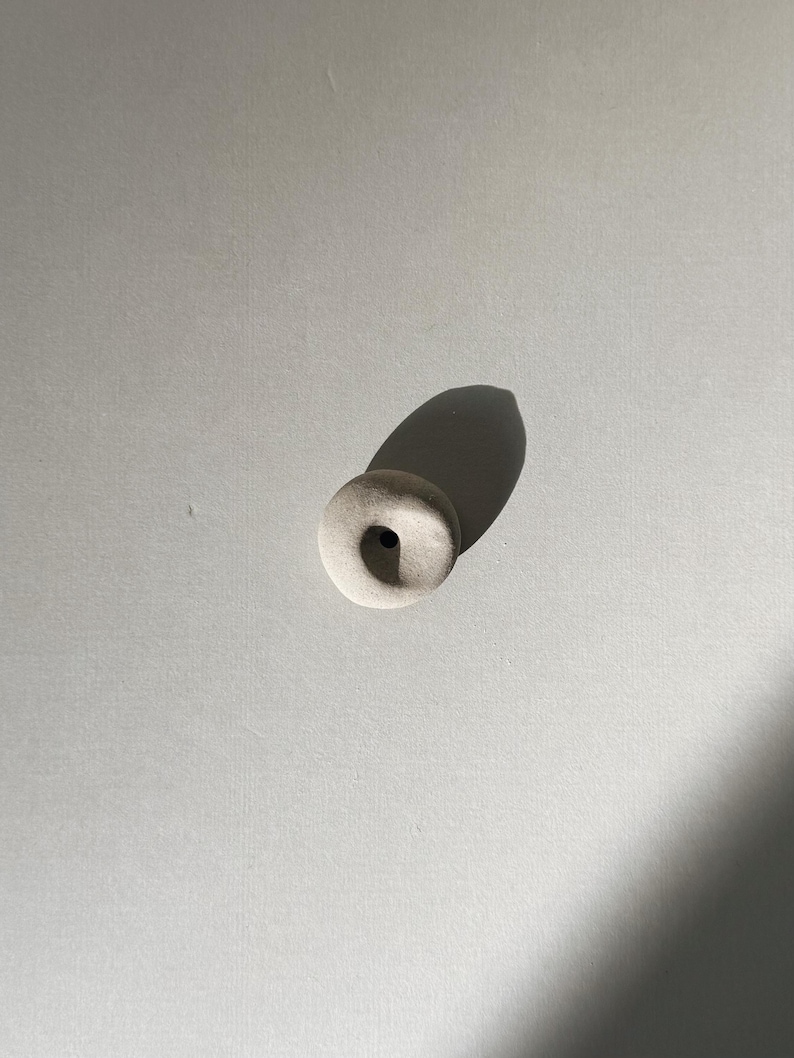 Puede incluir: Una peque&ntilde;a cuenta de cer&aacute;mica blanquecina con forma circular y un agujero central. La cuenta es lisa y tiene un acabado mate. La imagen est&aacute; tomada desde arriba, con una sombra proyectada hacia un lado.