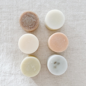 May include: Six round bars of soap in various colors and textures. The soaps are arranged in two rows of three on a light beige fabric background.