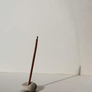 Puede incluir: Un palito de incienso marr&oacute;n se coloca en un peque&ntilde;o soporte de piedra gris claro. El soporte es redondo con un peque&ntilde;o agujero para sujetar el incienso. El fondo es una superficie blanca lisa.