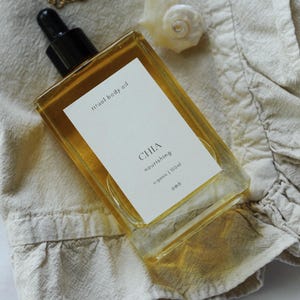 May include: A rectangular glass bottle of "ritual body oil" with a black dropper top, labeled "CHIA nourishing organic 100ml". A gold chain necklace with a seashell pendant rests on a beige fabric. The oil is amber-colored.