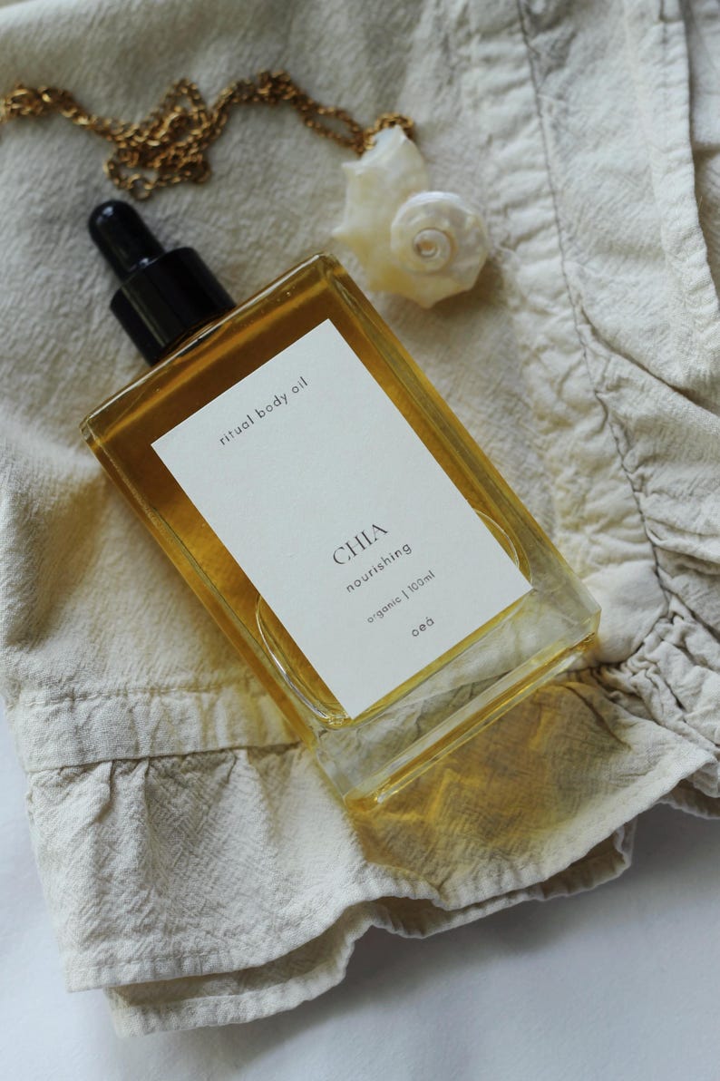 May include: A rectangular glass bottle of "ritual body oil" with a black dropper top, labeled "CHIA nourishing organic 100ml". A gold chain necklace with a seashell pendant rests on a beige fabric. The oil is amber-colored.