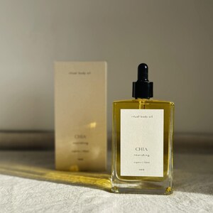 May include: A clear glass bottle of "ritual body oil" with a black dropper top, next to a matching box. The bottle has a white label with the word "CHIA" and "nourishing" printed on it. The oil appears golden in color.