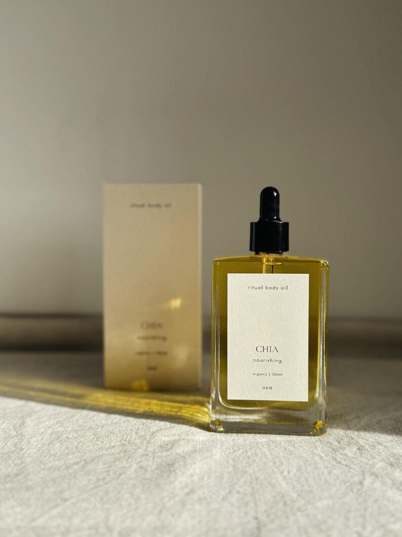 May include: A clear glass bottle of "ritual body oil" with a black dropper top, next to a matching box. The bottle has a white label with the word "CHIA" and "nourishing" printed on it. The oil appears golden in color.
