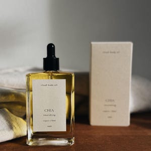 May include: A rectangular glass bottle of "ritual body oil" with a black dropper top, next to a matching box. The bottle has a white label with the word "CHIA" and other text. The oil is a golden color.