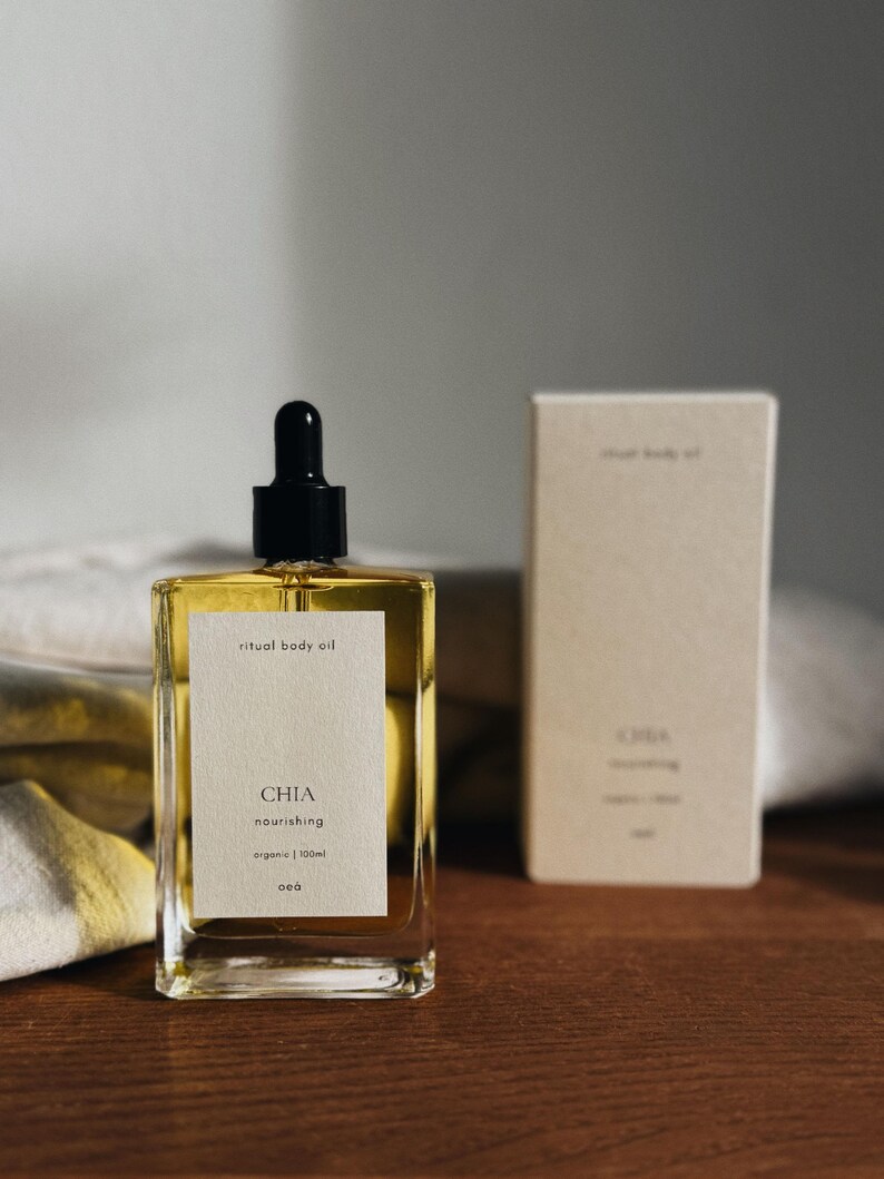 May include: A rectangular glass bottle of "ritual body oil" with a black dropper top, next to a matching box. The bottle has a white label with the word "CHIA" and other text. The oil is a golden color.