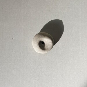 Puede incluir: Un peque&ntilde;o objeto redondo de cer&aacute;mica blanquecina con un agujero central. El objeto proyecta una larga sombra sobre una superficie gris clara. La pieza tiene una est&eacute;tica minimalista, lo que sugiere un art&iacute;culo decorativo o funcional.