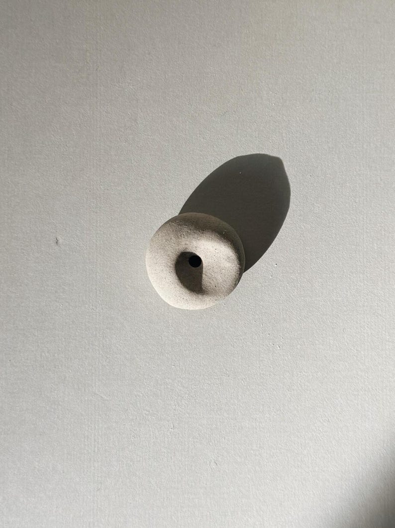 Puede incluir: Un peque&ntilde;o objeto redondo de cer&aacute;mica blanquecina con un agujero central. El objeto proyecta una larga sombra sobre una superficie gris clara. La pieza tiene una est&eacute;tica minimalista, lo que sugiere un art&iacute;culo decorativo o funcional.