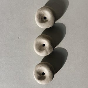 Puede incluir: Tres objetos circulares de color gris claro con un agujero central est&aacute;n dispuestos verticalmente. Cada objeto proyecta una larga sombra oscura sobre la superficie gris claro. Los objetos tienen un aspecto texturizado.