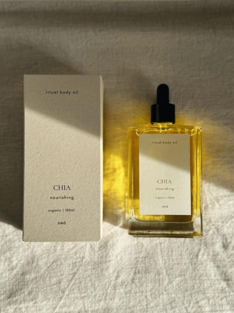 May include: A rectangular glass bottle of ritual body oil with a black dropper cap, next to a matching box. The bottle and box are labeled with the text "CHIA nourishing organic | 100ml". The oil is amber-colored.