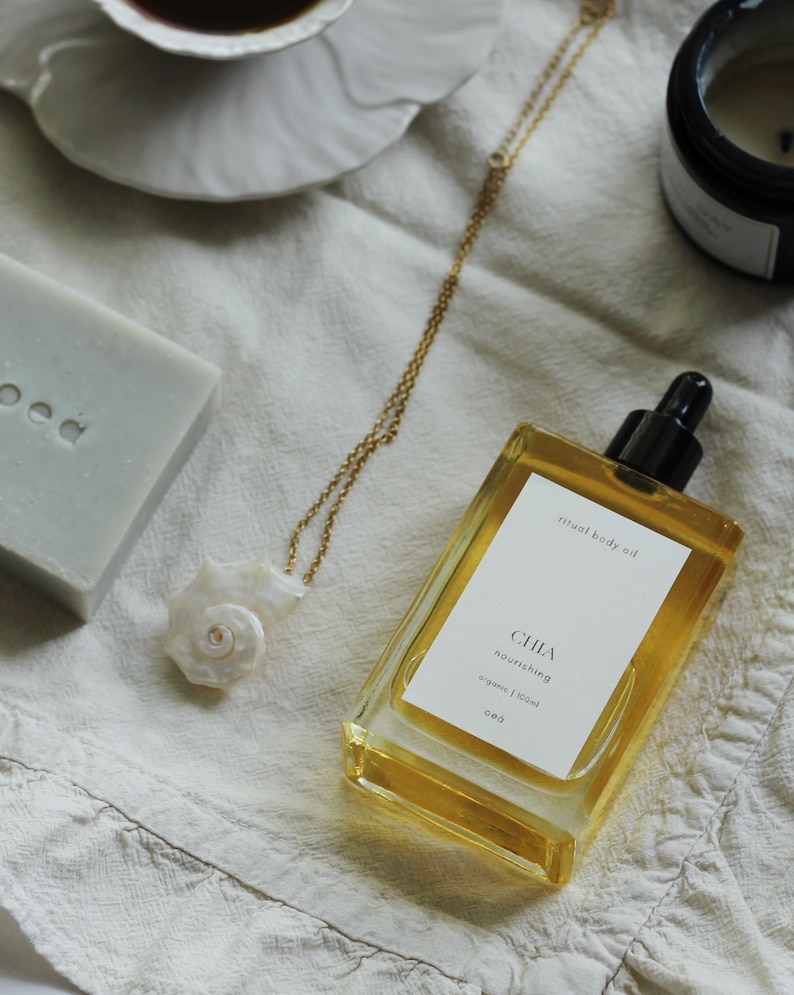 May include: A gold chain necklace with a white seashell pendant rests on a white cloth. A rectangular bottle of body oil with a black dropper and a white label is next to the necklace. A bar of soap and a cup of coffee are also in the image.