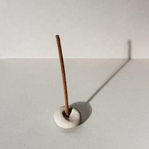 Puede incluir: Un peque&ntilde;o porta incienso de cer&aacute;mica blanquecina con una varilla de incienso marr&oacute;n insertada. El soporte es circular con un agujero en el centro. La varilla de incienso est&aacute; de pie, proyectando una sombra.
