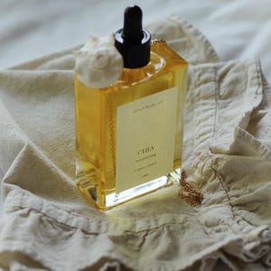 May include: A clear, rectangular bottle of "Chia nourishing" body oil with a black dropper and a white seashell accent. The bottle sits on a beige cloth, with a gold chain and a label that reads "organic | soul".
