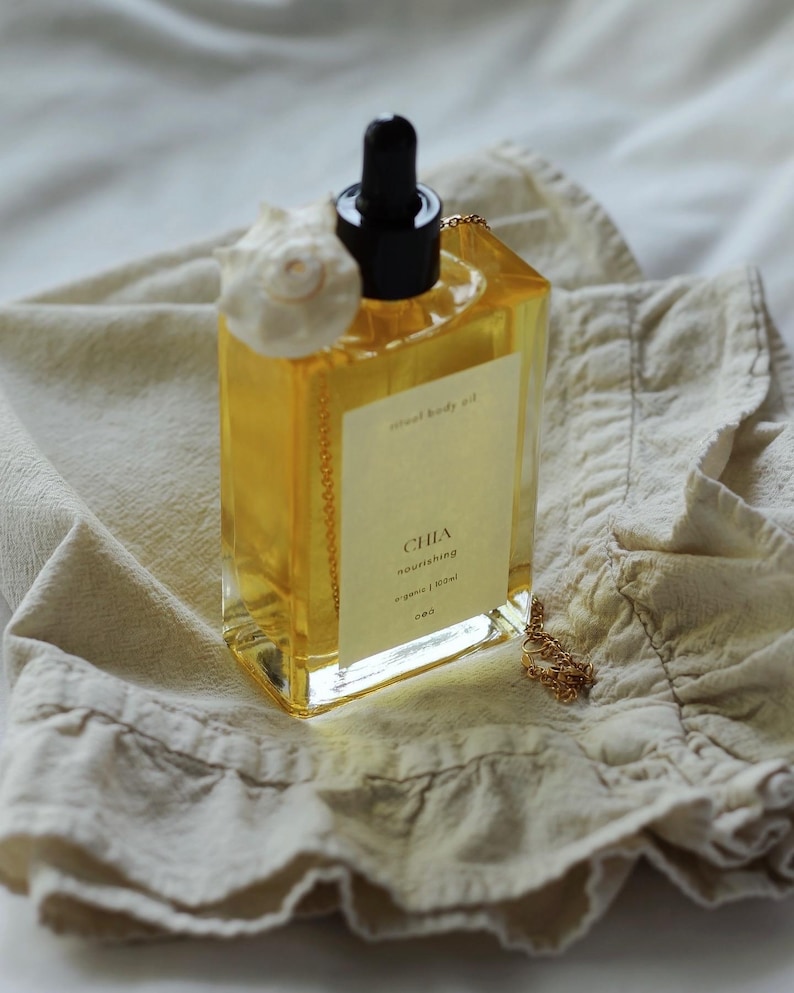 May include: A clear, rectangular bottle of "Chia nourishing" body oil with a black dropper and a white seashell accent. The bottle sits on a beige cloth, with a gold chain and a label that reads "organic | soul".
