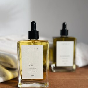 May include: Two clear glass bottles of ritual body oil with black droppers. The front bottle has a white label with the word "CHIA" and the words "nourishing" and "organic | 100ml". The oil inside is yellow.