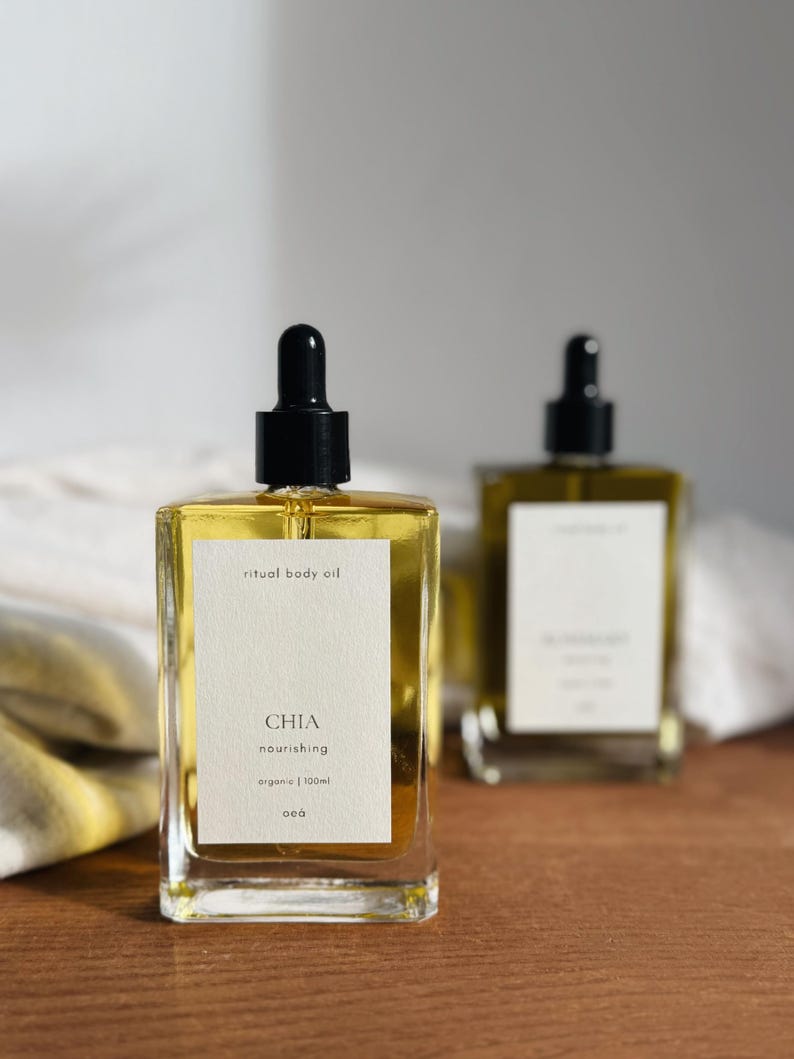 May include: Two clear glass bottles of ritual body oil with black droppers. The front bottle has a white label with the word "CHIA" and the words "nourishing" and "organic | 100ml". The oil inside is yellow.