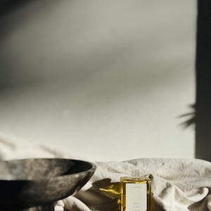 May include: A rectangular glass bottle of body oil with a white label that reads "Chia". The bottle is filled with a golden liquid and sits on a wooden surface. A dark dropper and a decorative bowl are also present.