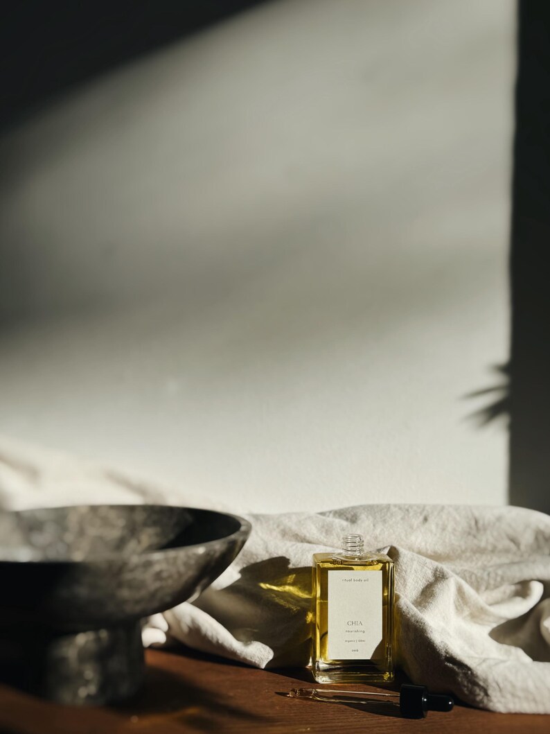 May include: A rectangular glass bottle of body oil with a white label that reads "Chia". The bottle is filled with a golden liquid and sits on a wooden surface. A dark dropper and a decorative bowl are also present.