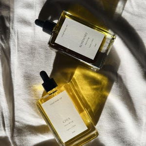 May include: Two rectangular glass bottles of ritual body oil with black droppers. One bottle is labeled "Rosemary detoxing" and the other "Chia nourishing". The bottles are filled with a golden liquid and sit on a white surface.