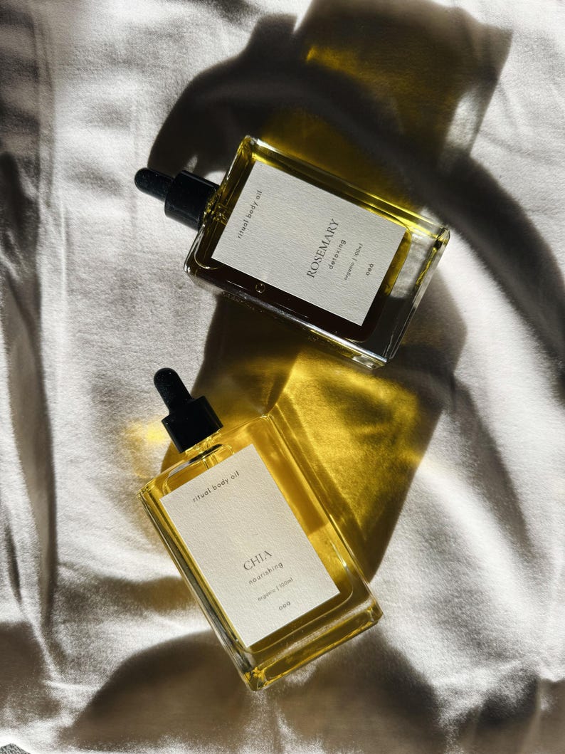 May include: Two rectangular glass bottles of ritual body oil with black droppers. One bottle is labeled "Rosemary detoxing" and the other "Chia nourishing". The bottles are filled with a golden liquid and sit on a white surface.