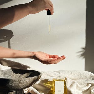 May include: A clear glass bottle of oil with a black dropper is held above an open hand. The bottle is rectangular with a gold label that reads "Chia Nourishing Face Oil". A marble bowl and white cloth are in the background.