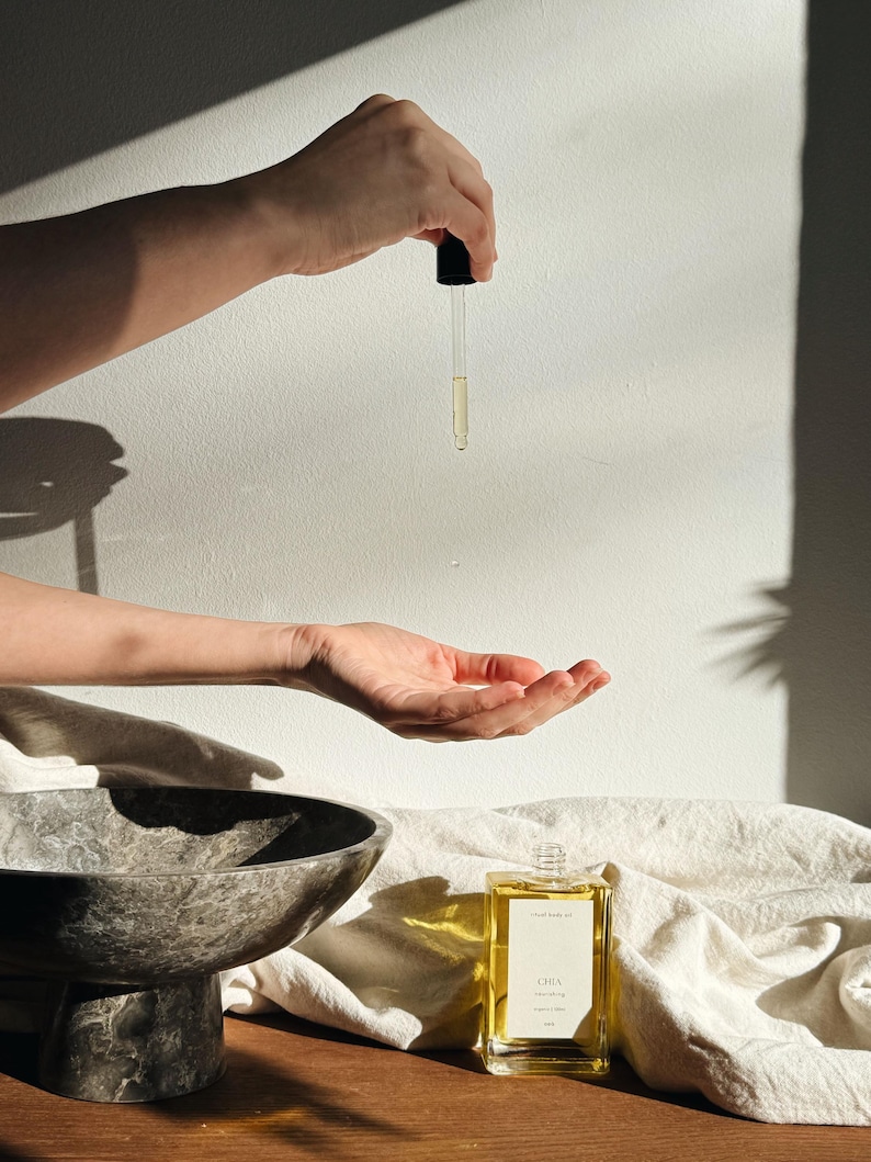May include: A clear glass bottle of oil with a black dropper is held above an open hand. The bottle is rectangular with a gold label that reads "Chia Nourishing Face Oil". A marble bowl and white cloth are in the background.