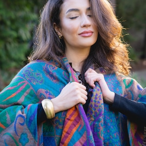 May include: A vibrant paisley patterned shawl in teal, purple, orange, and pink. The shawl is draped over the shoulders. A gold cuff bracelet is visible on the left wrist. The background is a blurred outdoor scene.