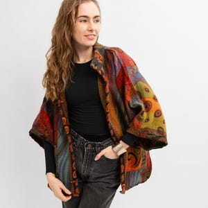 May include: A colorful patchwork jacket with a mix of patterns and colors, including red, orange, and blue. The jacket has wide sleeves and is worn over a black long-sleeve shirt and dark jeans. A silver bracelet is visible.