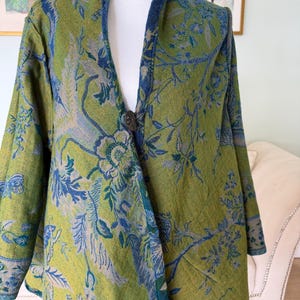 Könnte beinhalten: Eine langärmelige Jacke in einem leuchtenden Olivgrün mit einem detaillierten Webmuster aus blauen Blumen- und Pflanzenmotiven. Das Kleidungsstück hat einen einzelnen dunklen Knopfverschluss in der vorderen Mitte und einen strukturierten Kragen mit blauem Saum.
