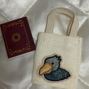以下が含まれることがあります： 大きな黄色い嘴と青い羽を持つハシビロコウの刺繍ワッペンが付いたオフホワイトのキャンバス地ミニトートバッグと、表紙に金色の太陽の紋章が描かれた小さな長方形のマルーン色の本。