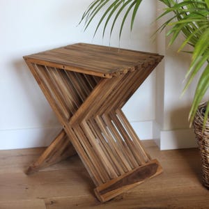 Può includere: Uno sgabello pieghevole in legno con finitura naturale. Lo sgabello ha un telaio a forma di X e una seduta a doghe. Lo sgabello è alto circa 46 cm e largo 36 cm. Una pianta verde in un cesto intrecciato è sullo sfondo.