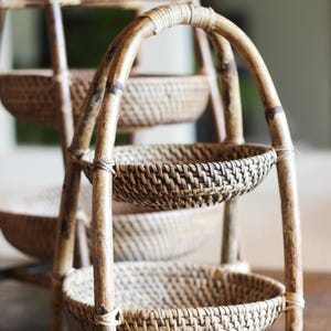 May include: A three-tiered bamboo basket stand with woven baskets. The stand has a curved handle and is made of natural-colored bamboo. The baskets are round and have a dark brown and tan woven pattern. The stand is on a wooden surface.