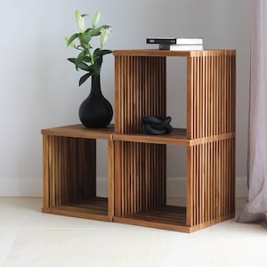 Puede incluir: Una estantería de madera con tres estantes. El estante superior tiene un libro titulado "NESS". El estante del medio tiene una escultura de cuerda negra. El estante inferior está vacío. La estantería está hecha de madera natural y tiene un diseño de listones.