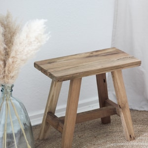 May include: A rustic wooden stool with a natural wood finish. The stool has a rectangular top and four legs. The stool is sitting on a woven rug.
