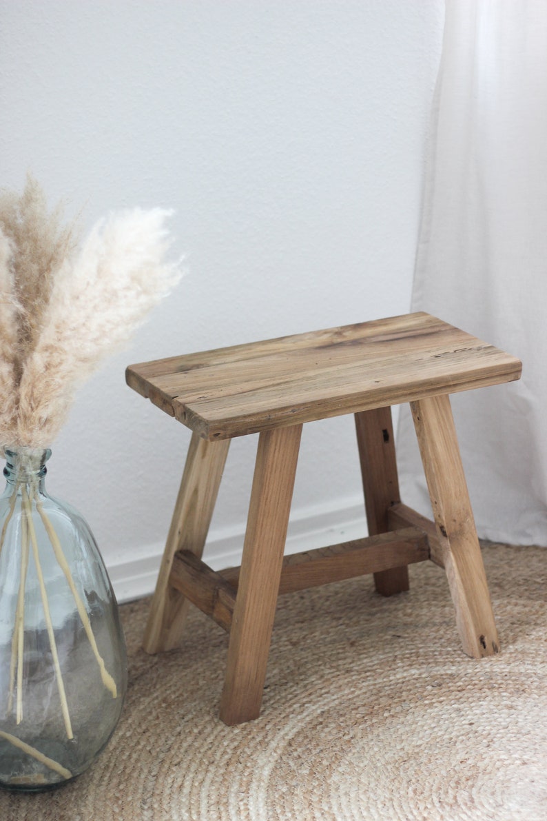 May include: A rustic wooden stool with a natural wood finish. The stool has a rectangular top and four legs. The stool is sitting on a woven rug.